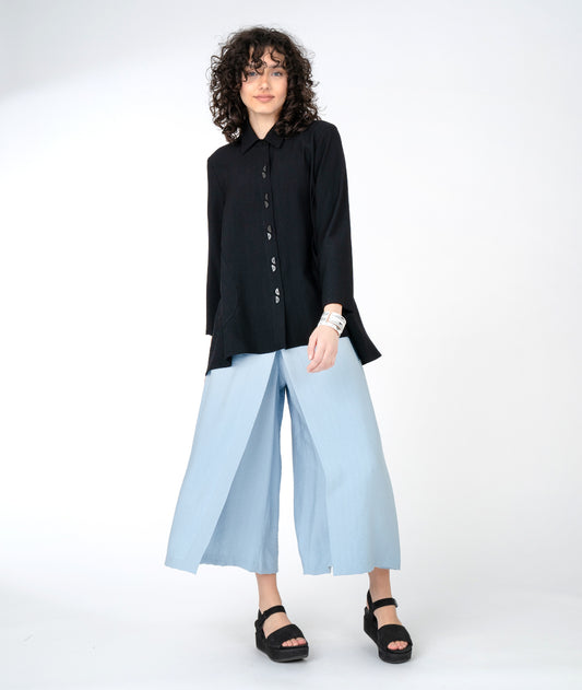 model in a wide leg blue pant with an overlapping front panel, worn with a black flowy full bodied button down blouse with large hip pockets