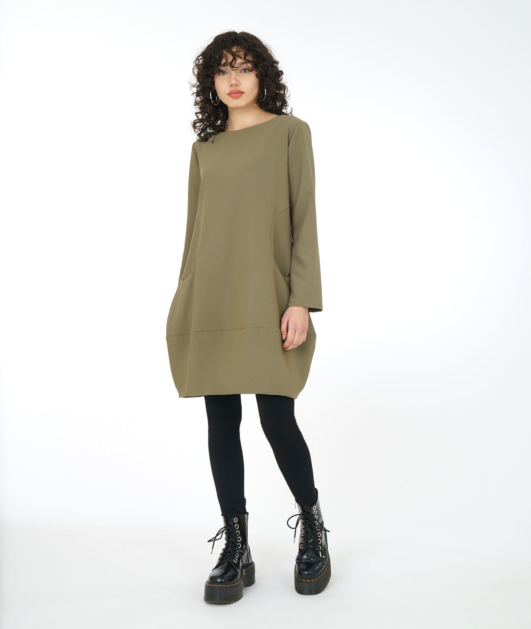 model in a sage green dress with long sleeves, a wide bottom hem, soft round neckline and curved side seams with a set in pocket on either side, worn with black leggings and boots