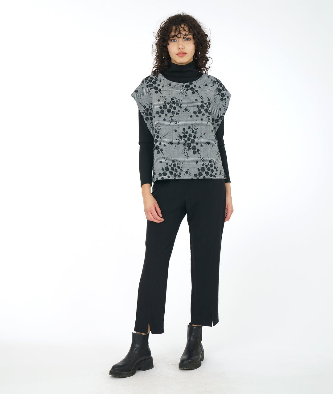 model in a slim leg black pant with a black turtleneck with a grey plaid poncho style vest layered on top. vest has a black pollen print