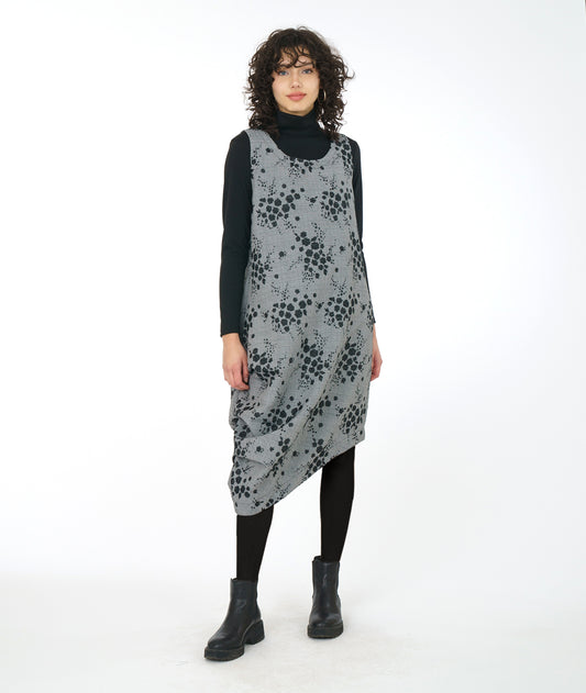 model in a grey plaid asymmetrical jumper dress with a black pollen print, worn over a black turtleneck, leggings and booties