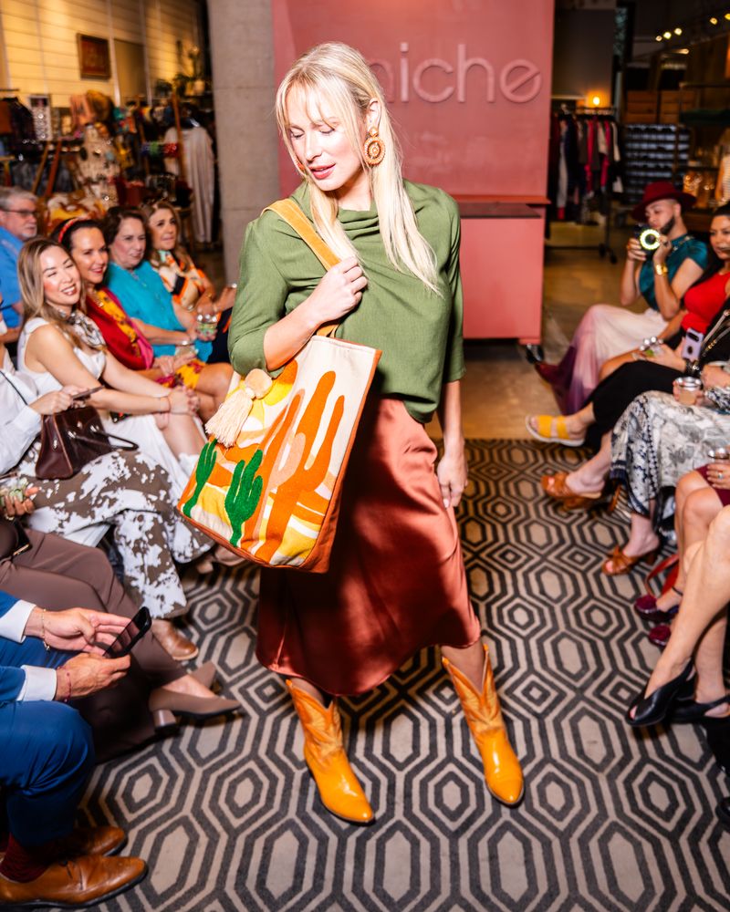 model posing on a runway wearing green cowl top, copper satin skirt, yellow cowboy boots and a desert scene tote bag