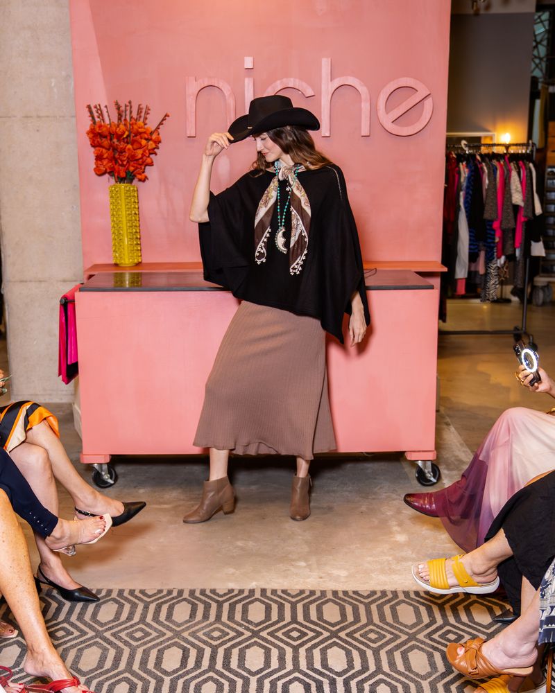 model wearing a black cowboy hat, black poncho, knit taupe skirt, brown scarf, long turquoise beaded moon necklace and boots walking down a runway