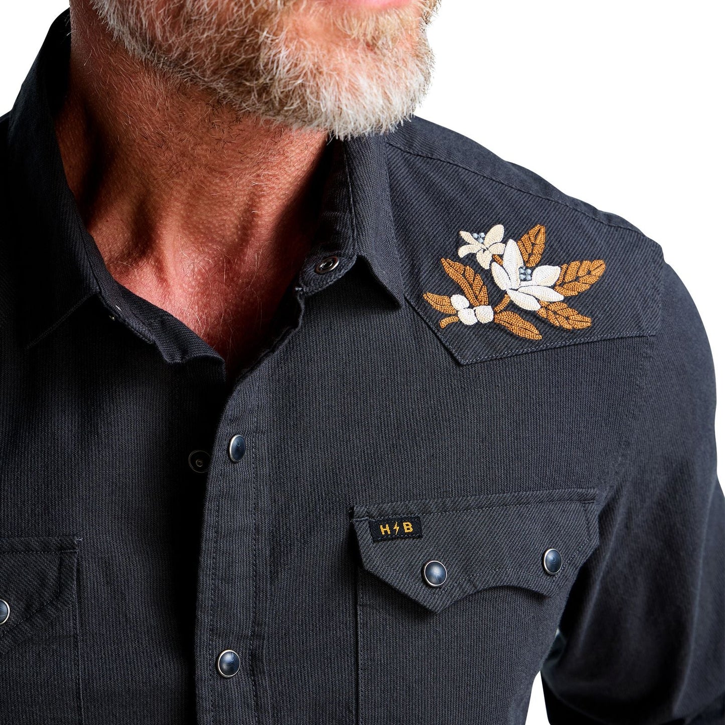 Close-up of a man wearing a black western-style button-up shirt with a floral embroidered patch on the shoulder and a small HB logo on the chest pocket.