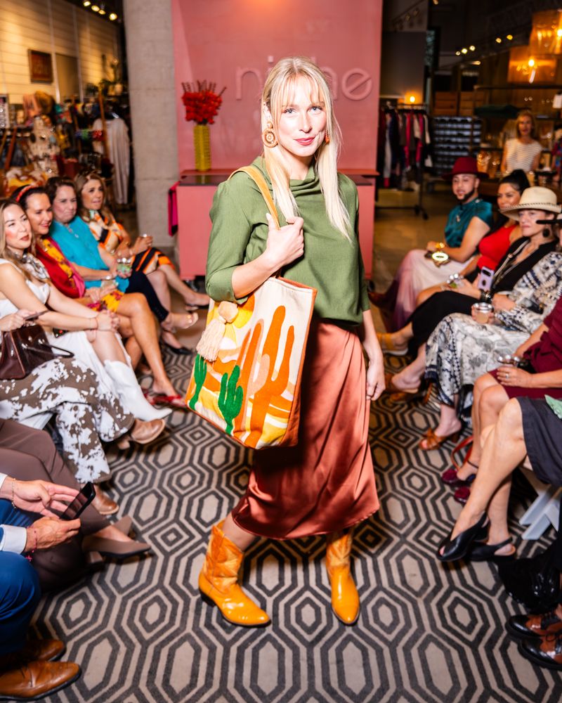 model posing on a runway wearing green cowl top, copper satin skirt, yellow cowboy boots and a desert scene tote bag