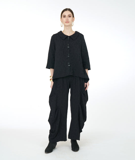 model in a wide leg black pant with exaggerated tucks up the sides, worn with a matching black jacket with a dipped hem at the sides and 3/4 sleeves