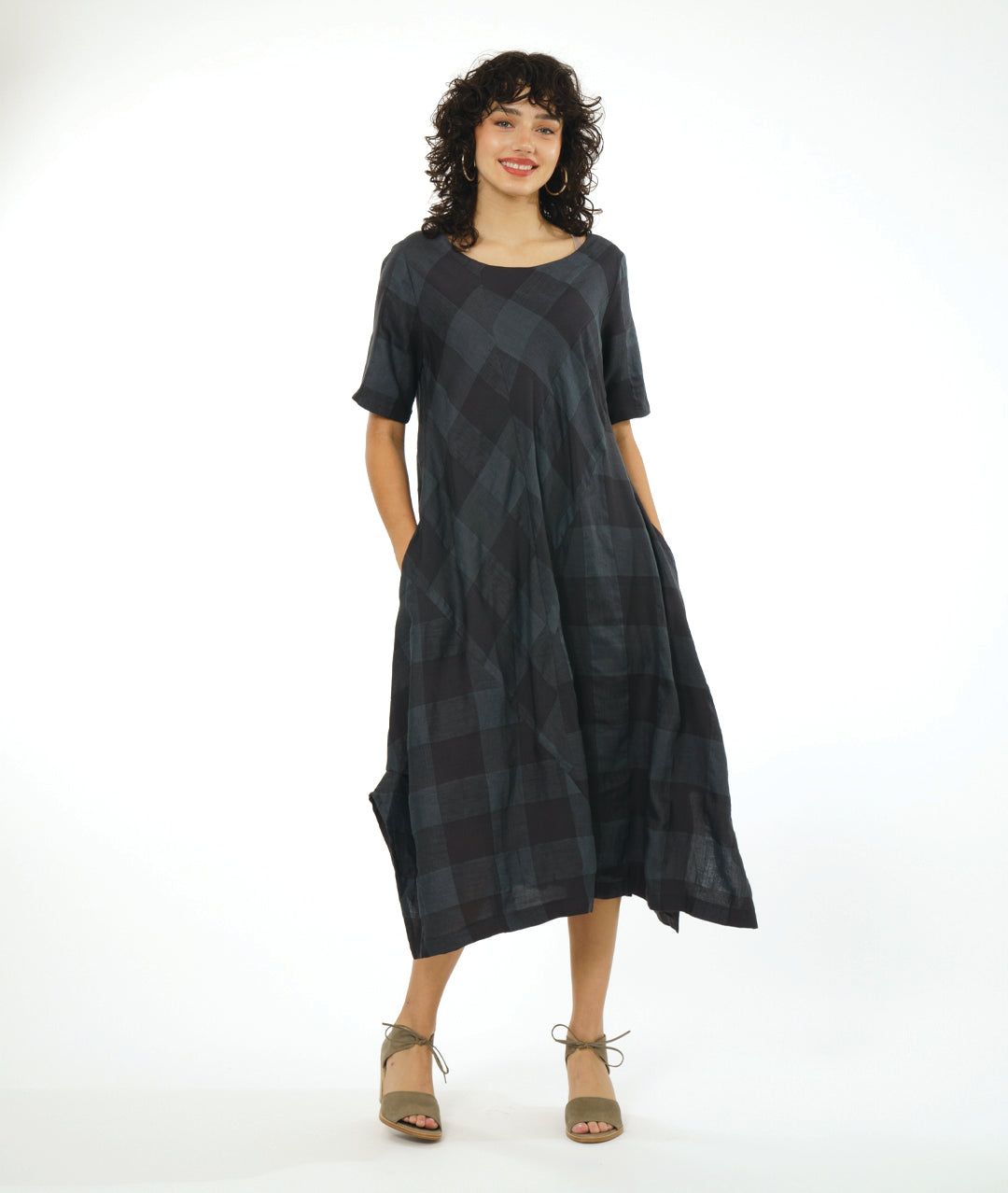 model in a midi length dress with short sleeves, a round neckline and tucks at the sides. dress has a large grid print on shades of deep gray with multi directional panels