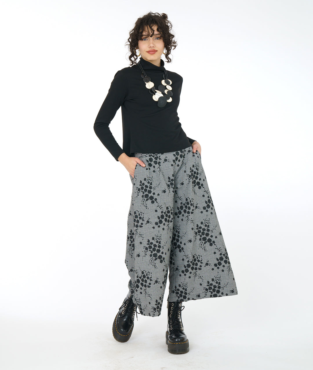 model in a black turtleneck and oversized chunky black and white necklace, worn with a cropped wide leg grey plaid pant with a black pollen print