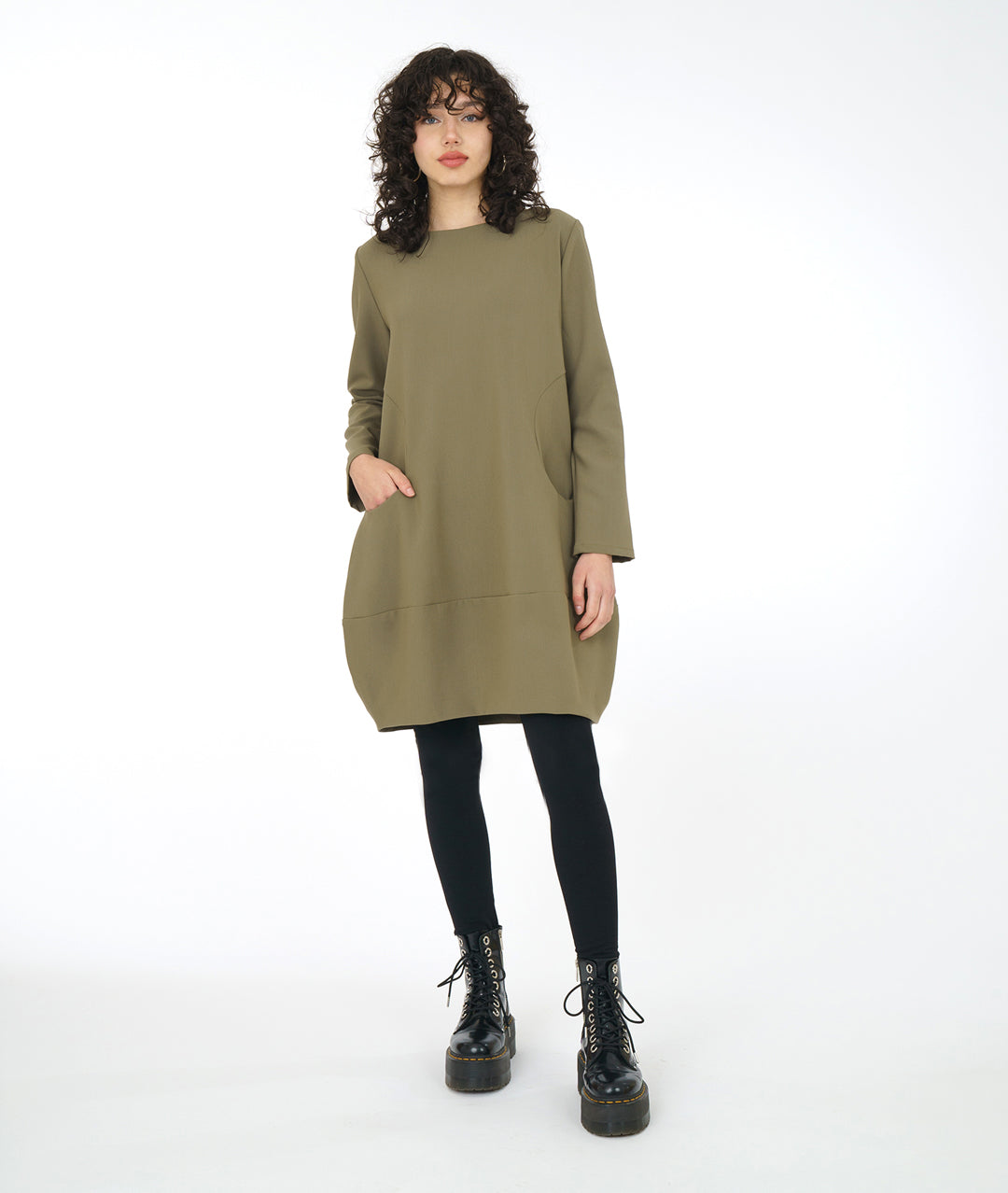 model in a sage green dress with long sleeves, a wide bottom hem, soft round neckline and curved side seams with a set in pocket on either side, worn with black leggings and boots
