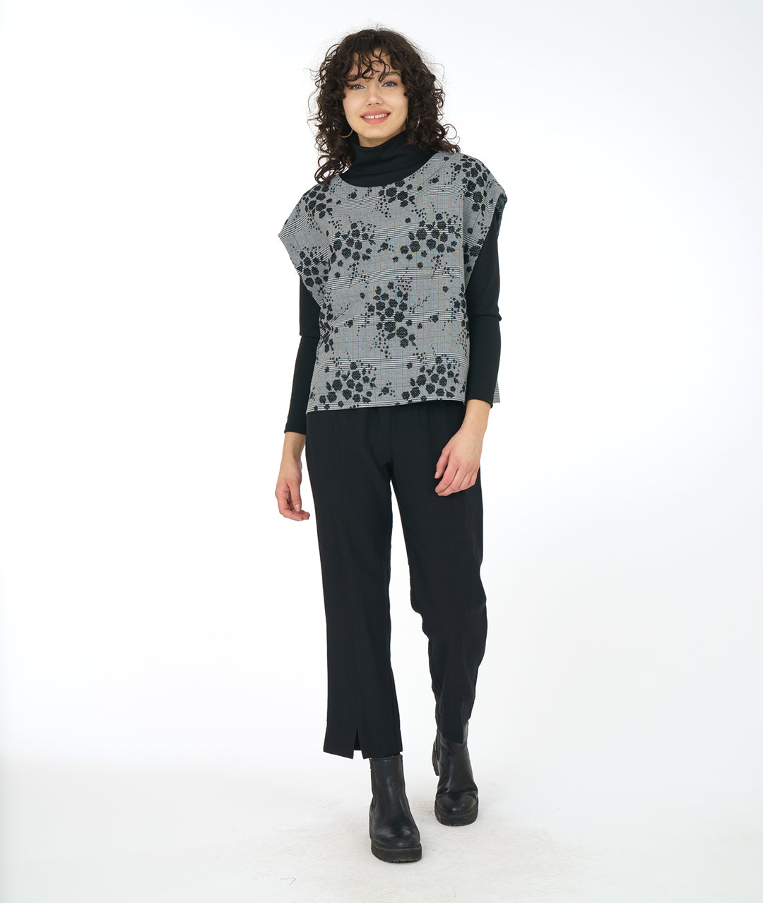 model in a slim leg black pant with a black turtleneck with a grey plaid poncho style vest layered on top. vest has a black pollen print