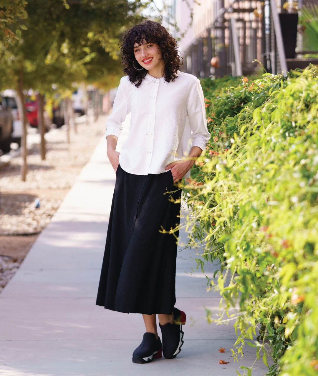 model in a black wide leg pant with overlapping apron panels in the front, worn with a white button down blouse with a twin button detail and 3/4 sleeves