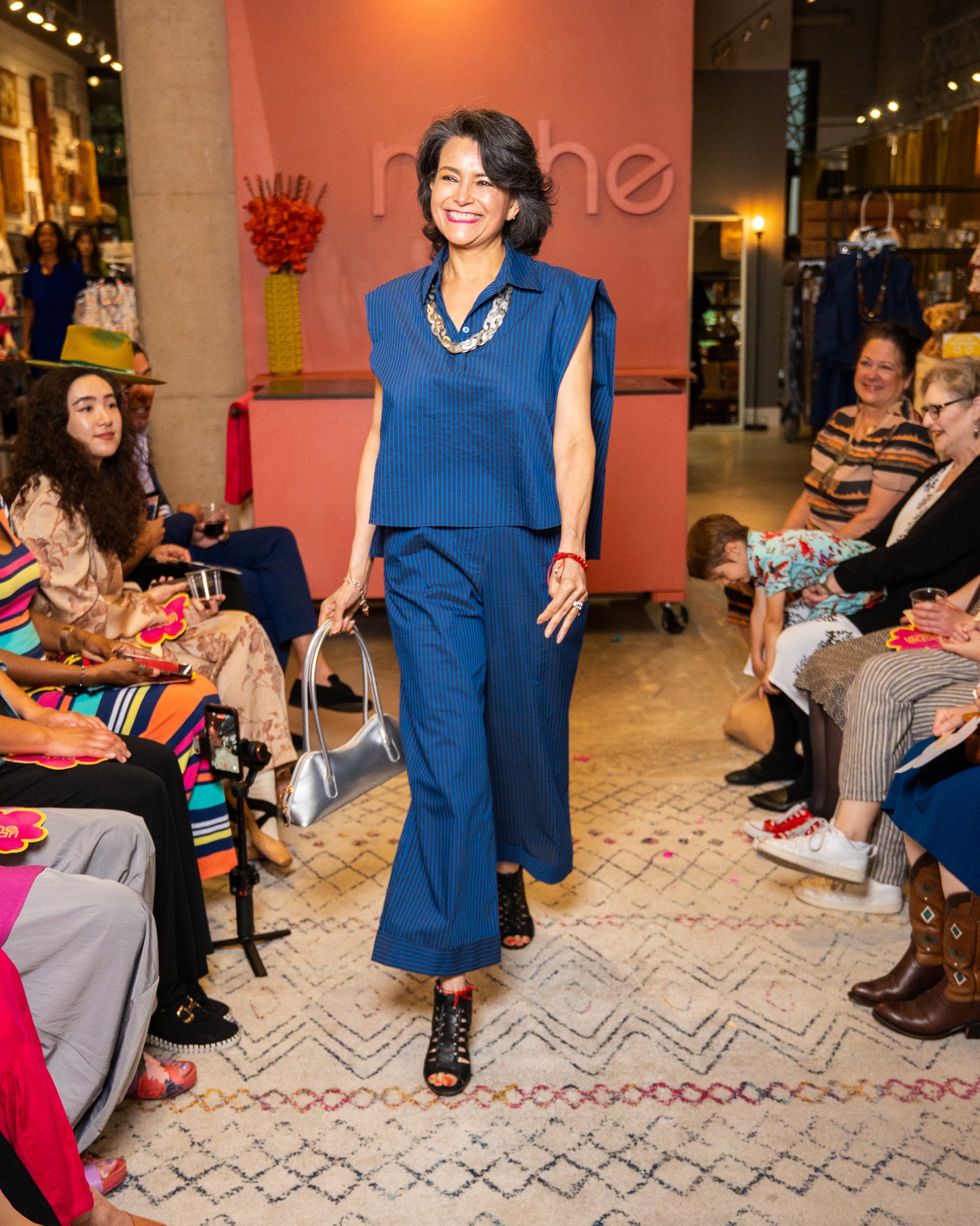 Woman in a blue top and pant and silver necklace and purse walking down a runway at a fashion event