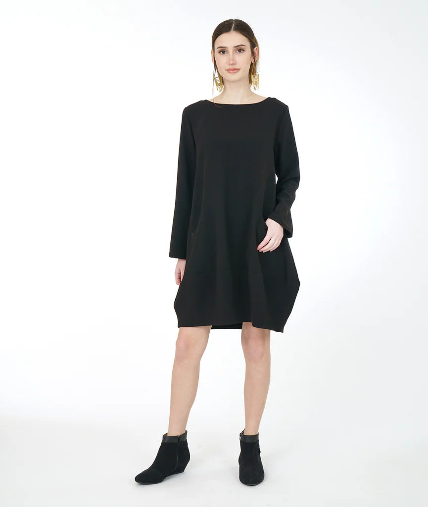 model in a black dress with long sleeves, a wide bottom hem, soft round neckline and curved side seams with a set in pocket on either side, worn with black shoes.