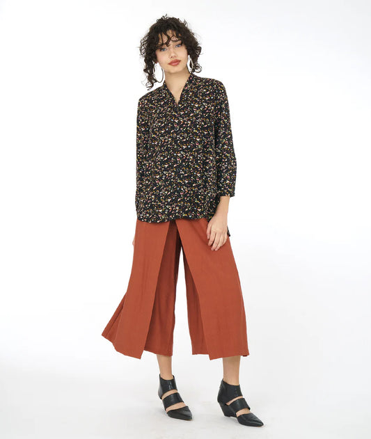 model wearing black terrazzo print button down blouse with long sleeves and a flowy, full back. top has a button at each wrist and a deep neckline with a standing collar. paired with dark orange pants and black shoes.