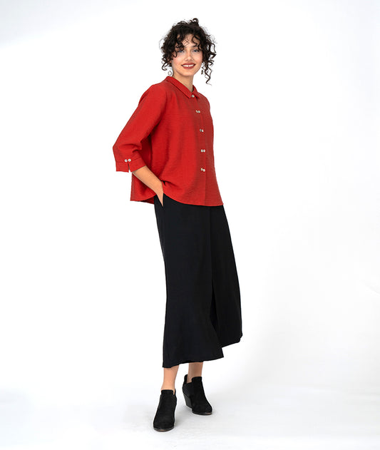 model in a wide leg black pant with an apron overlay, worn with a red boxy button down blouse with 3/4 sleeves and a twin button detail