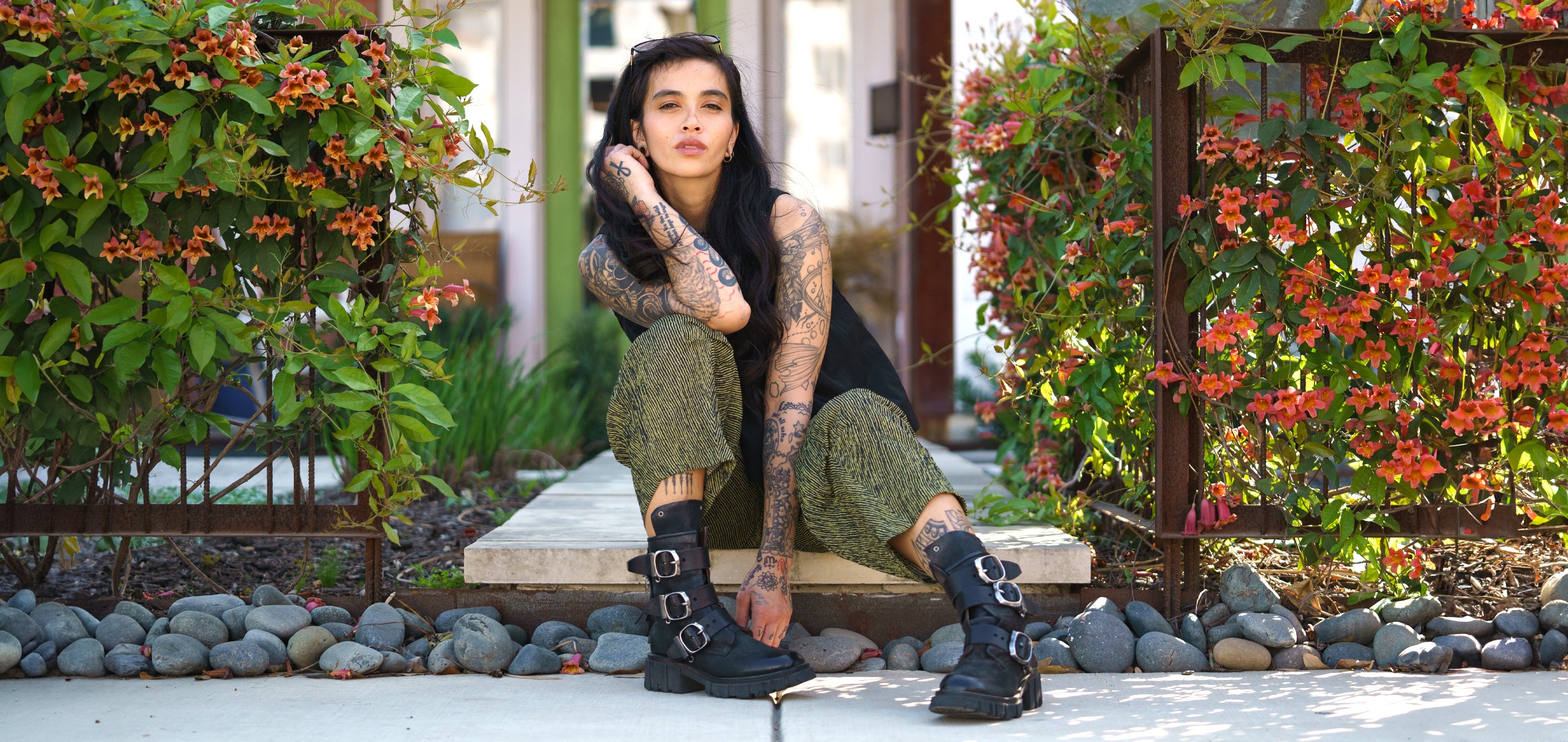 model sitting on a sidewalk in a black boots and top, with a black and green striped pant, surrounded by flowers