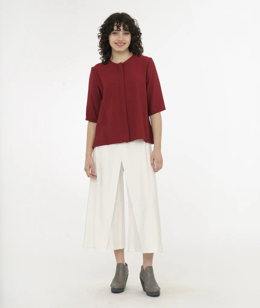 model wearing brick red top with a hidden buttoned placket, 3/4 sleeves, and a round neckline worn with white pants.