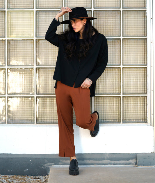 model in a rust color straight cut pant, worn with a black pullover top with long sleeves and a stair step hem
