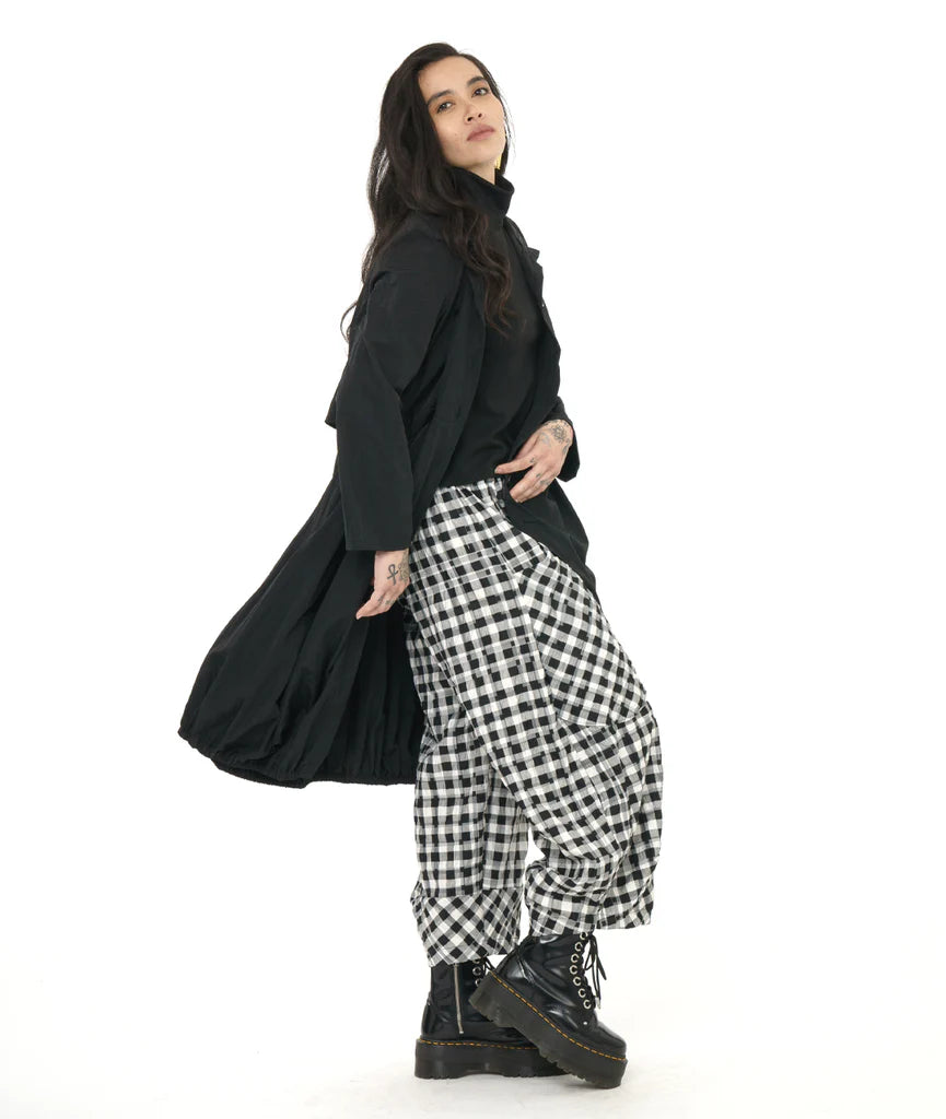 model wearing black lantern shape long jacket with cowl collar and round pockets with black top, plaid pants and boots.