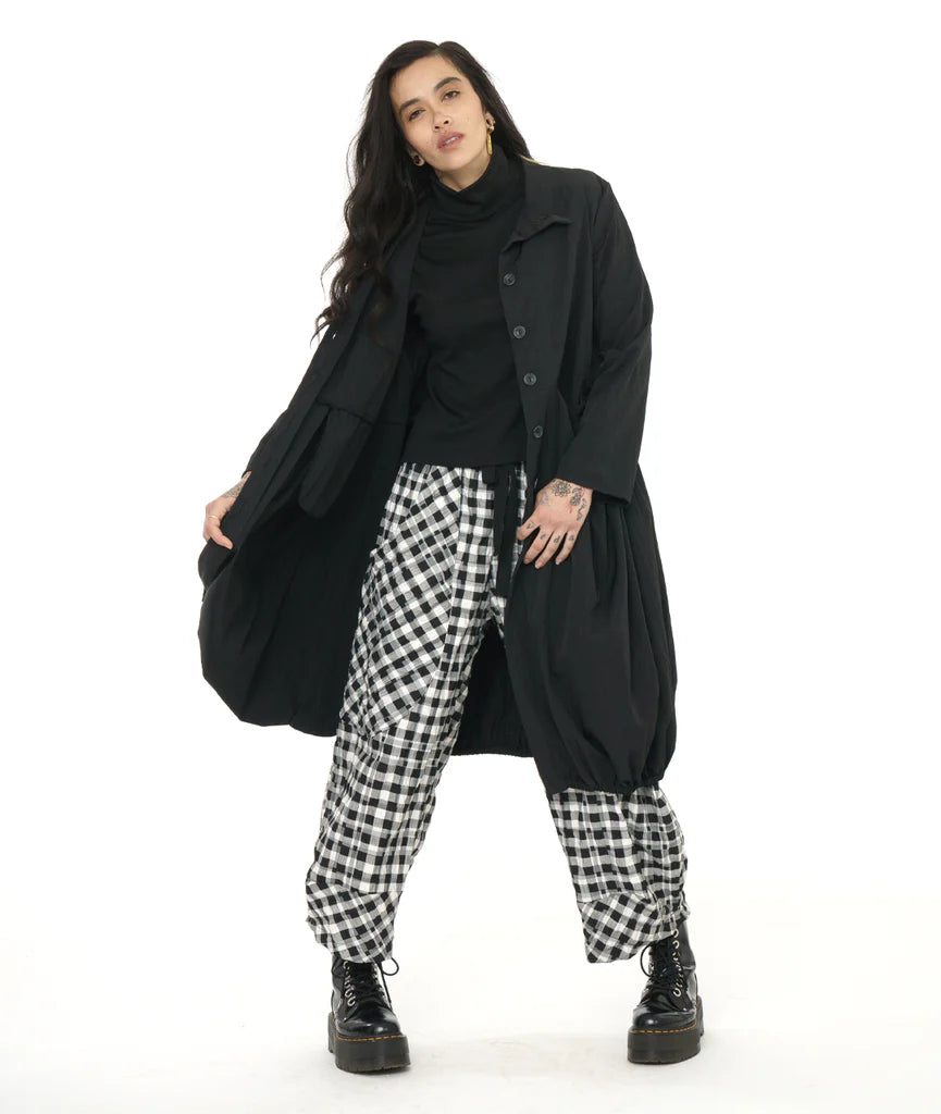 model wearing black lantern shape long jacket with cowl collar and round pockets with black top, plaid pants and boots.