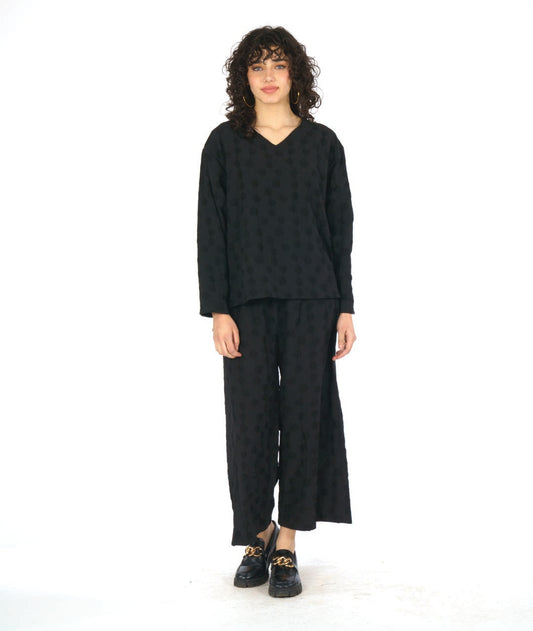 model in a black pullover blouse with a puckered dot texture. top has long sleeves, a v-neck and a stairstep hem. worn with a wide leg matching pant