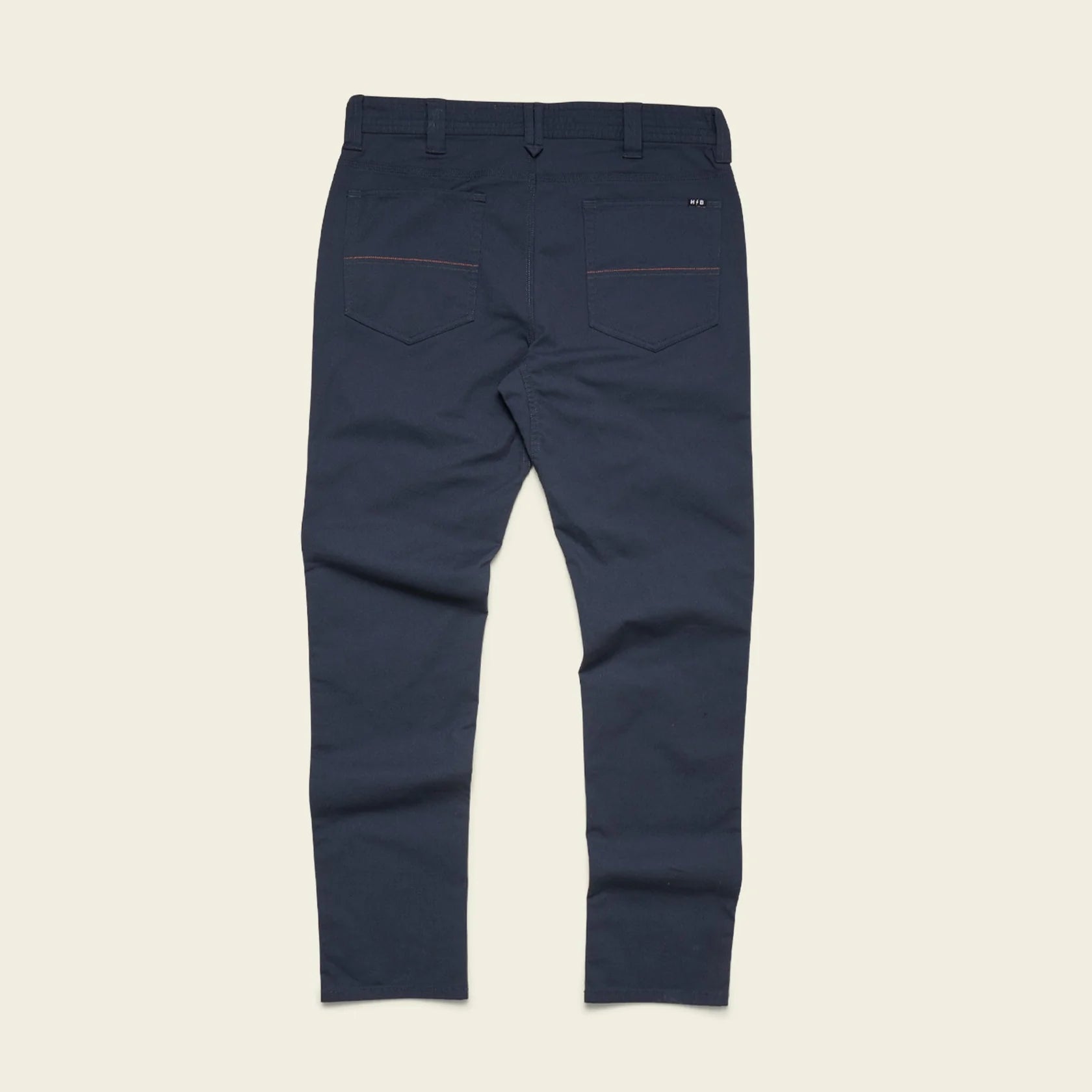 A pair of navy blue slim-fit trousers shown from the back, featuring two back pockets with thin red contrast stitching and a small white embroidered logo on the upper right pocket, laid flat against a light beige background.