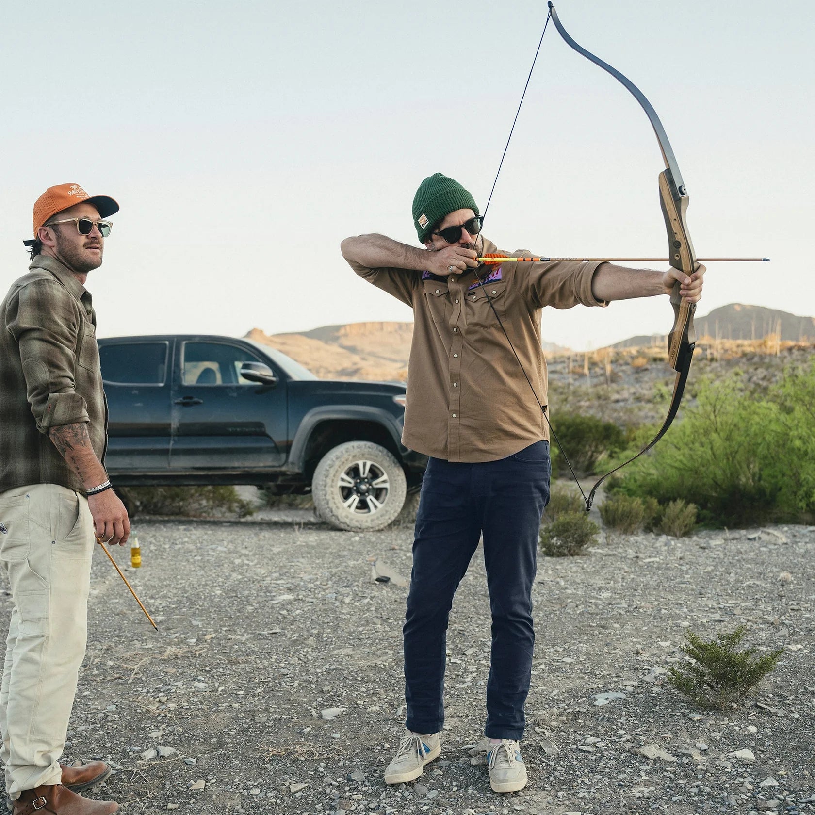 Two men in a desert setting with one aiming a bow and arrow and wearing navy pants with brown button up shirt.