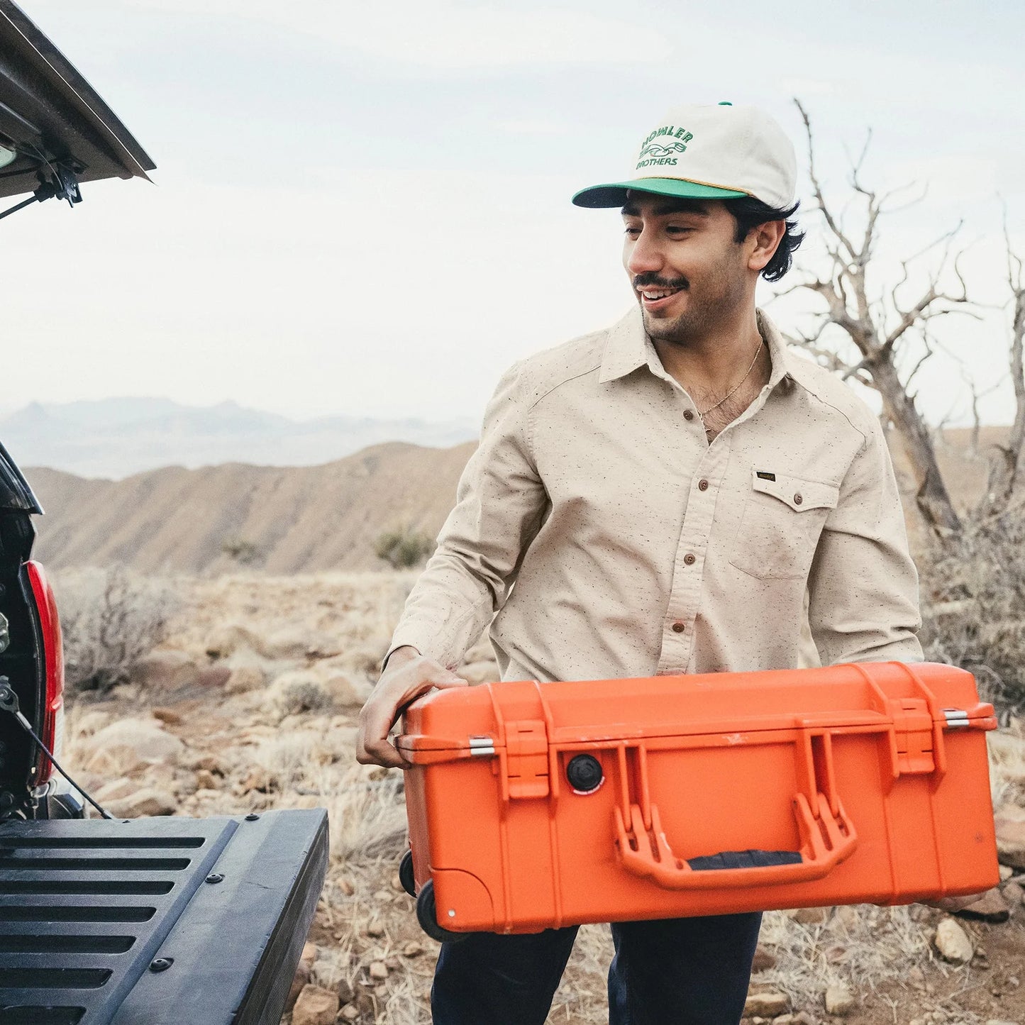 Man holding an orange suitcase in a desert-like setting with mountains in the background wearing beige flannel and hat.