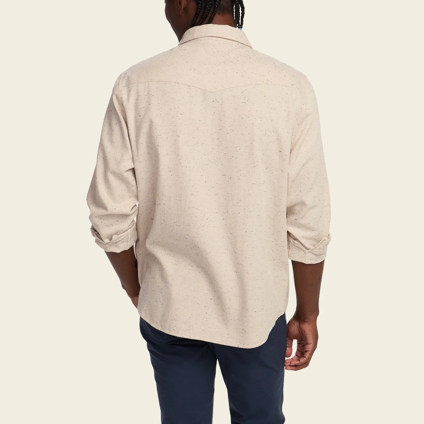 A man viewed from the back wearing a light beige speckled long-sleeve button-down shirt and navy pants, set against a solid off-white background.