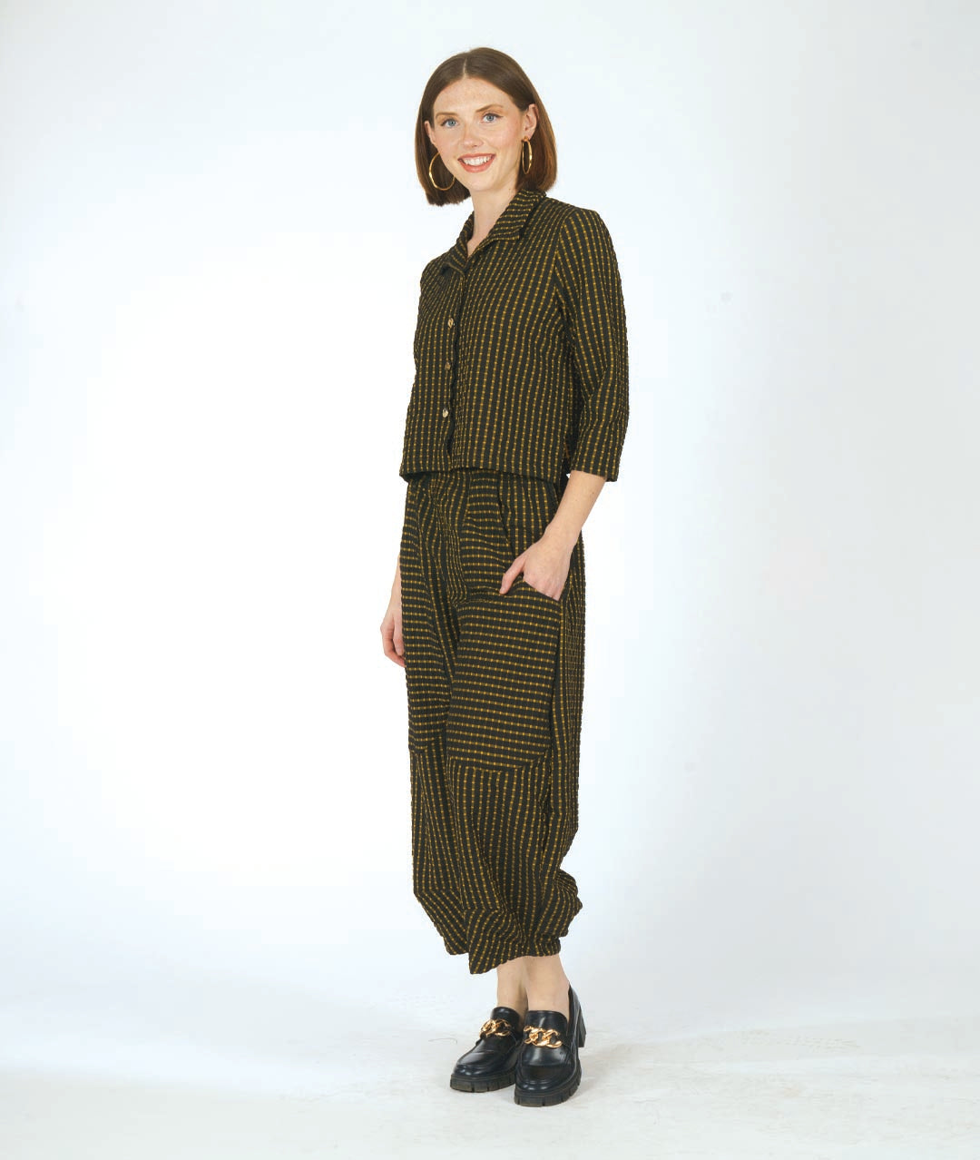 A woman modeling a matching two-piece set consisting of a cropped button-front collared shirt and wide-leg cropped pants, both in a small-scale black and yellow check pattern. She is wearing black loafers with gold hardware against a plain white studio background.