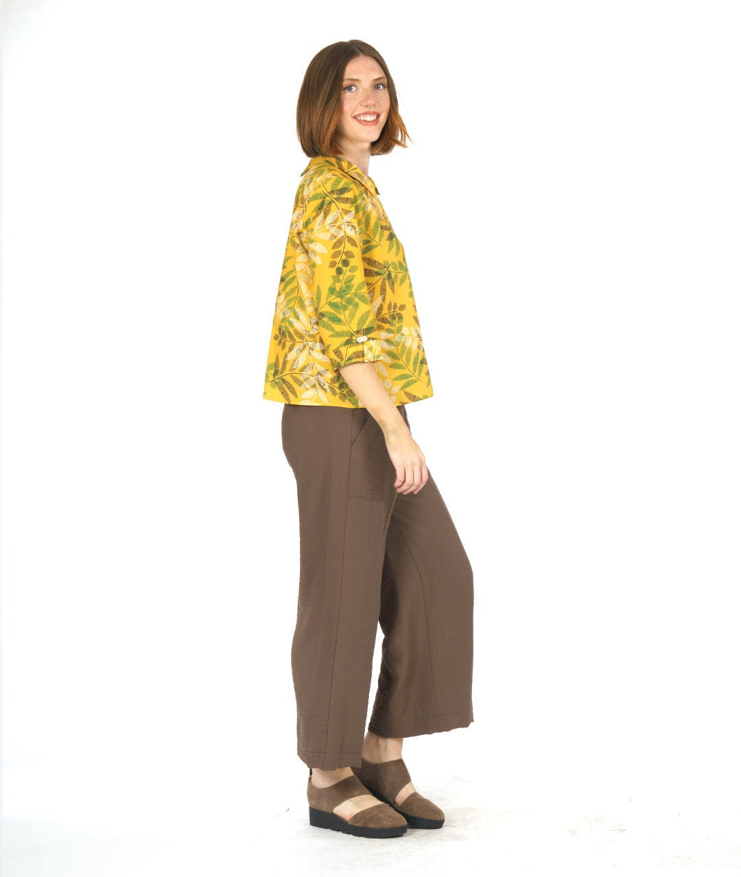model in a straight leg brown pant with a yellow leaf print button down blouse. blouse has 3/4 sleeves, a boxy body and a twin button detail at the sleeves, back and front center
