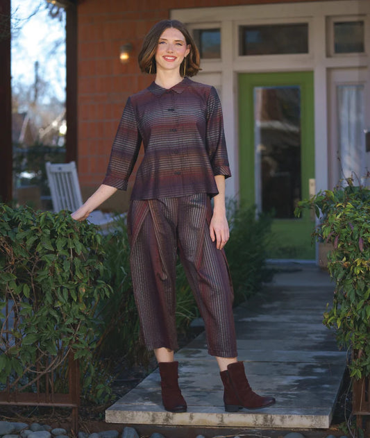 model wearing a matching button up 3/4 sleeve striped top and front pleated pants standing on a stone path with greenery and a building in the background