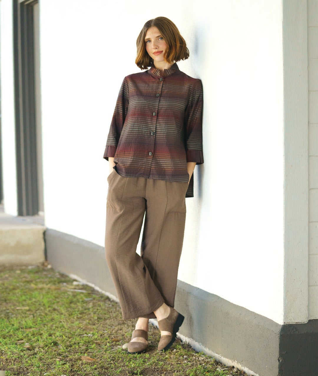 model in a red and brown tone striped blouse with 3/4 sleeves with a wide cuff and a mandarin style collar. worn with a straight leg brown pant