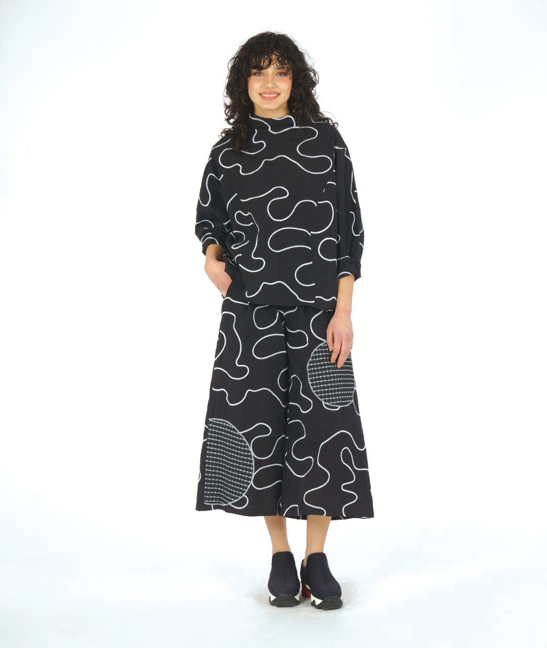 model in a black pant with a white swirl embroidery print and checkerboard patches. worn with a matching pullover top with princess seams and a cowl neckline