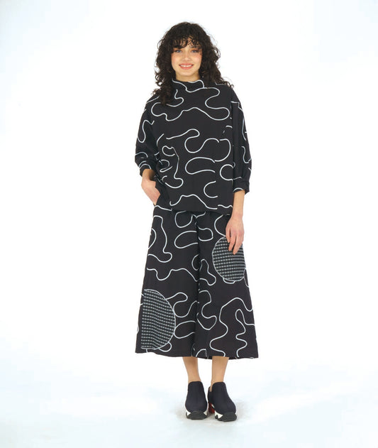 model in a black pant with a white swirl embroidery print and checkerboard patches. worn with a matching pullover top with princess seams and a cowl neckline