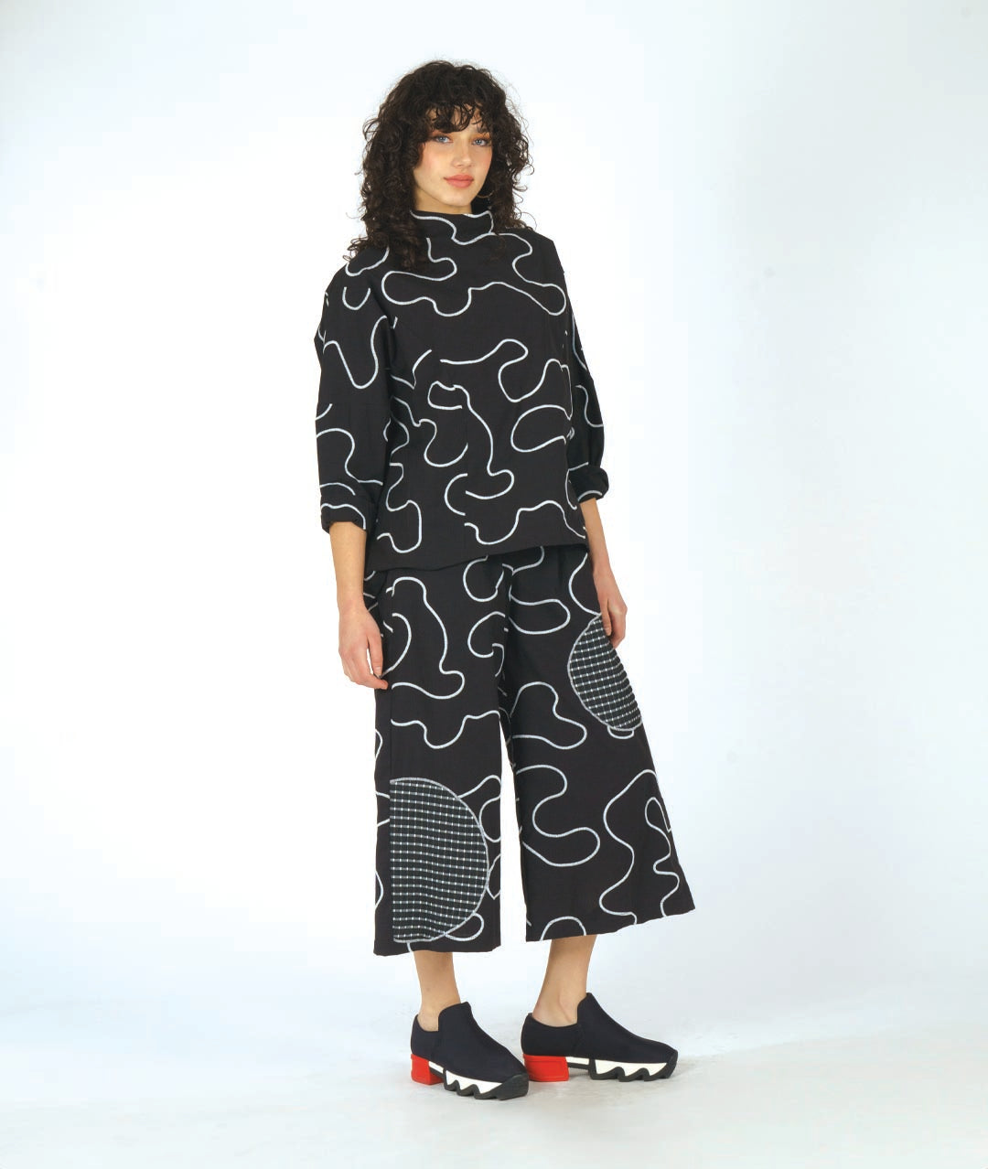 model in a black pant with a white swirl embroidery print and checkerboard patches. worn with a matching pullover top with princess seams and a cowl neckline