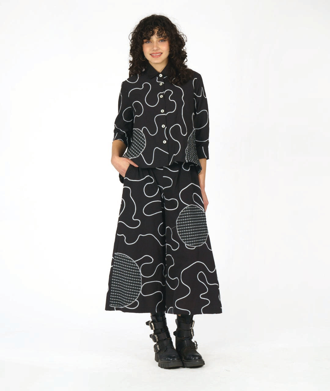 model wearing a black button down blouse with a bell shaped wide body. blouse has a white swirl print embroidered all over, and large round patches of check print fabric accented across. worn with a wide leg matching pant