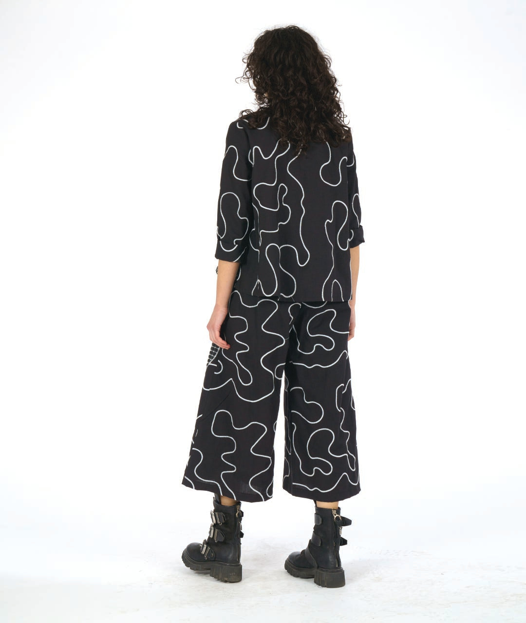 model wearing a black button down blouse with a bell shaped wide body. blouse has a white swirl print embroidered all over, and large round patches of check print fabric accented across. worn with a wide leg matching pant