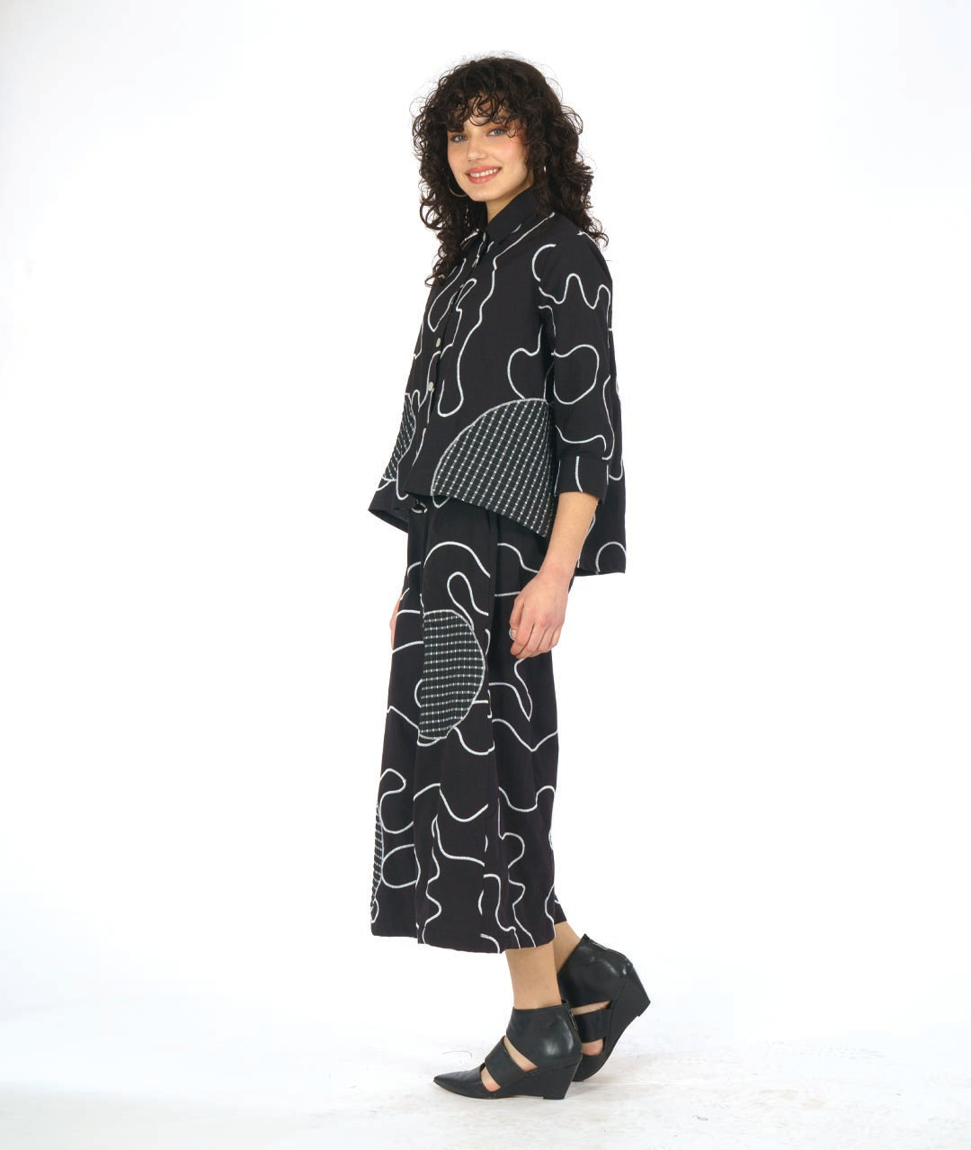 model wearing a black button down blouse with a bell shaped wide body. blouse has a white swirl print embroidered all over, and large round patches of check print fabric accented across. worn with a wide leg matching pant