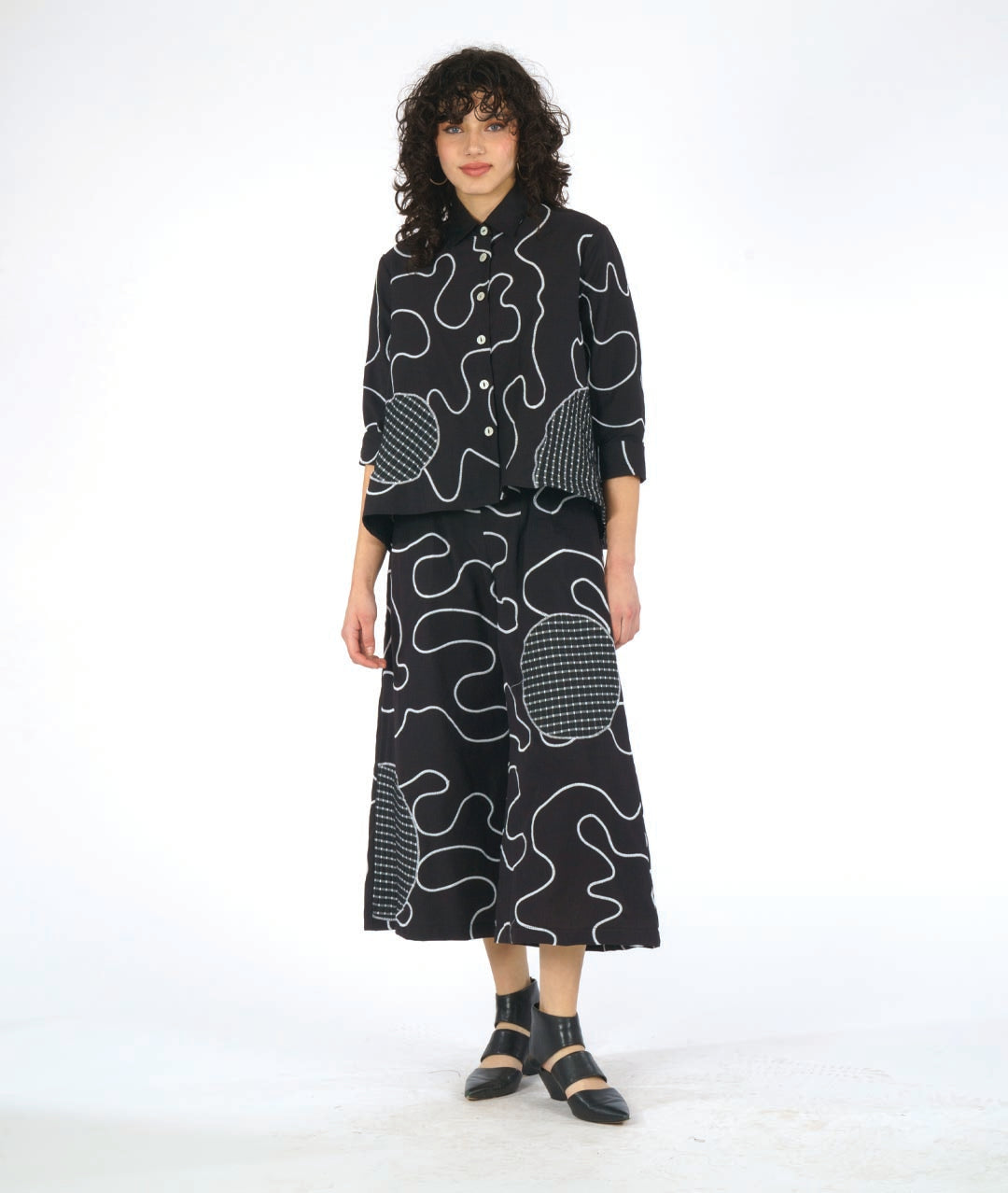 model wearing a black button down blouse with a bell shaped wide body. blouse has a white swirl print embroidered all over, and large round patches of check print fabric accented across. worn with a wide leg matching pant