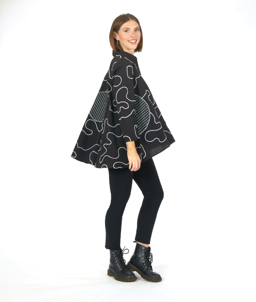 model in a black pant with a black button down blouse. blouse has a white swirl embroidery print and checkerboard patches
