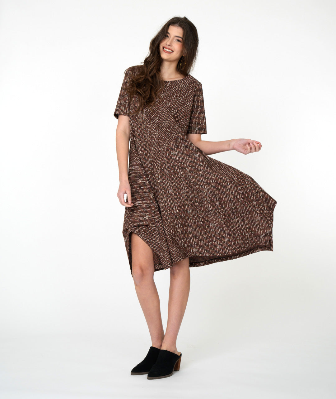 Woman wearing a brown patterned dress standing against a white brick wall.