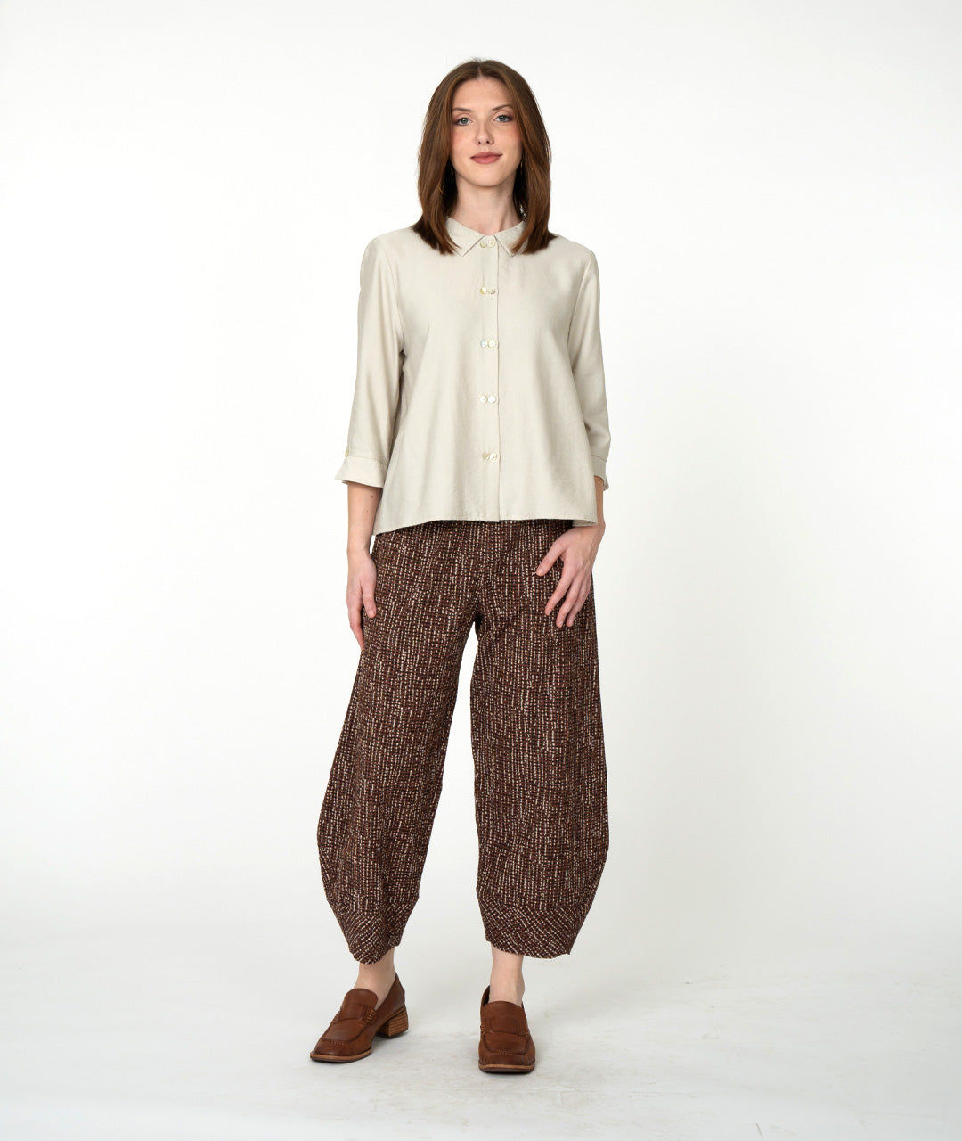 A woman stands against a plain white studio background wearing a light beige button-front blouse with three-quarter sleeves, brown patterned wide-leg cropped pants, and brown loafers.
