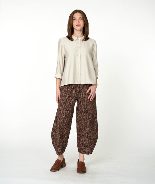 A woman stands against a plain white studio background wearing a light beige button-front blouse with three-quarter sleeves, brown patterned wide-leg cropped pants, and brown loafers.