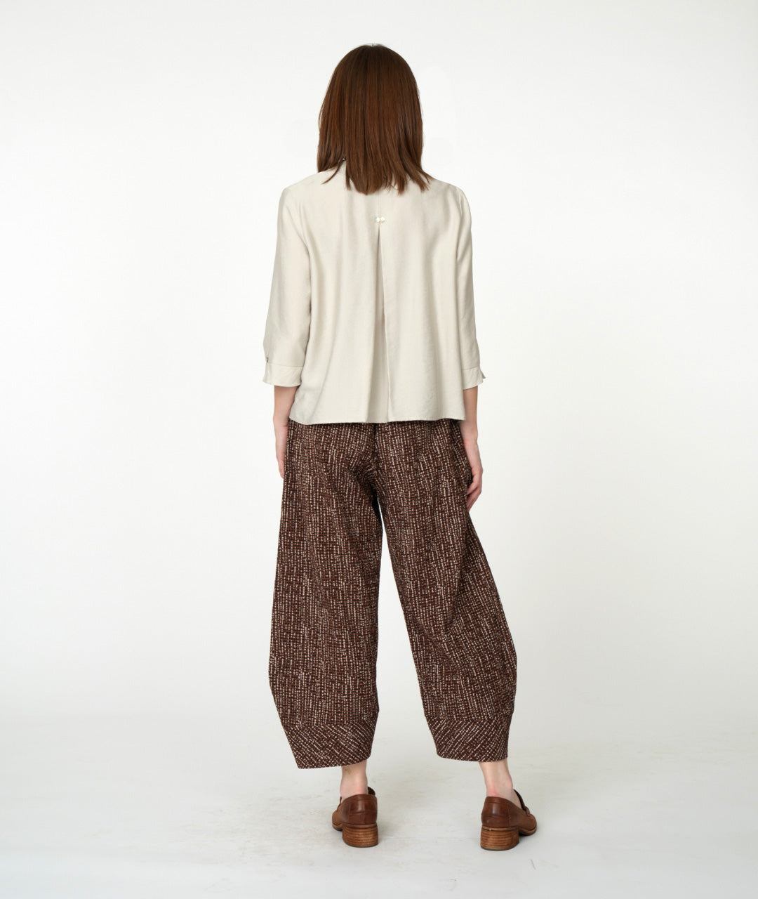 A woman stands against a plain white studio background wearing a light beige button-front blouse with three-quarter sleeves, brown patterned wide-leg cropped pants, and brown loafers.