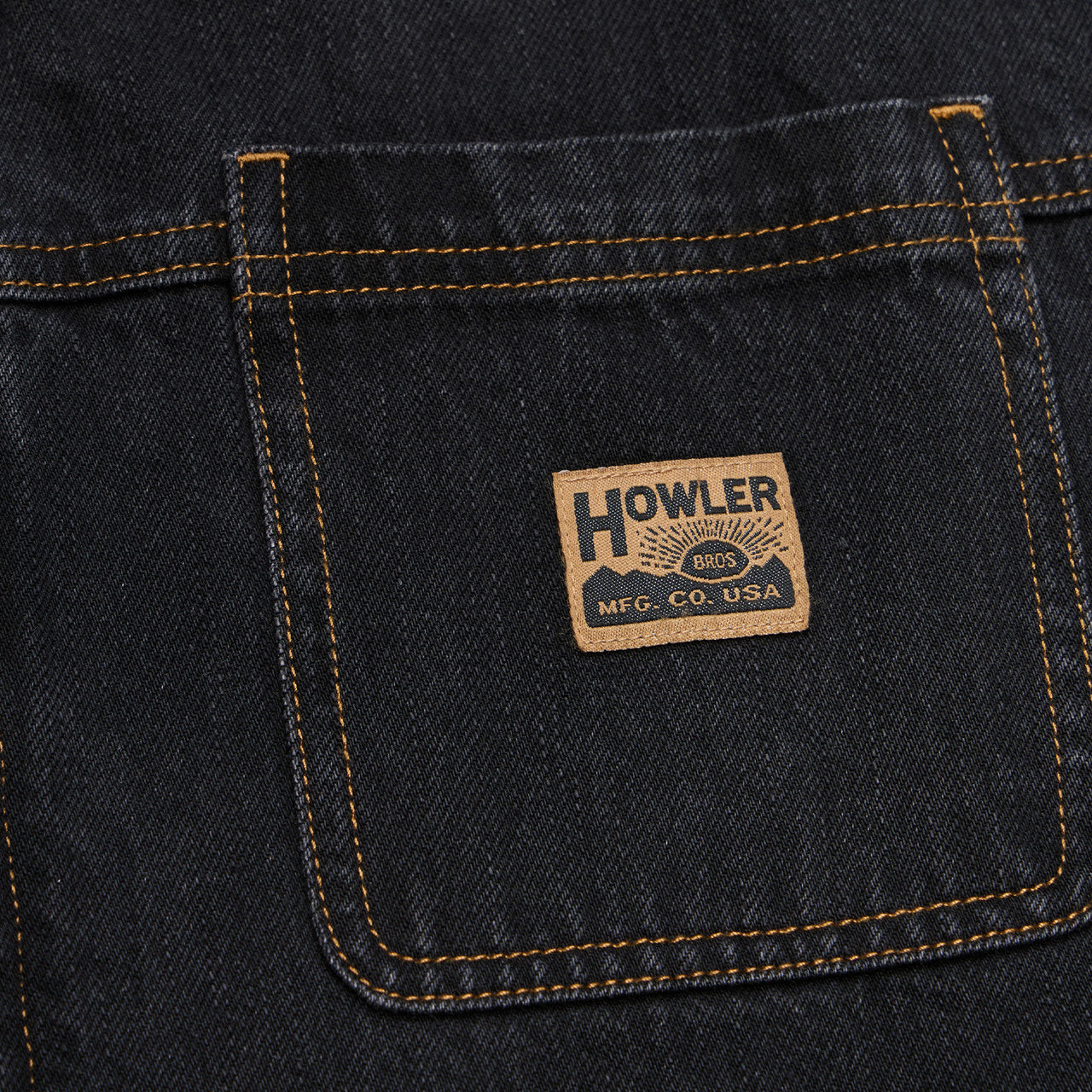 Close-up of a dark indigo denim back pocket with contrast tan stitching and a rectangular Howler Brand patch sewn onto the pocket.