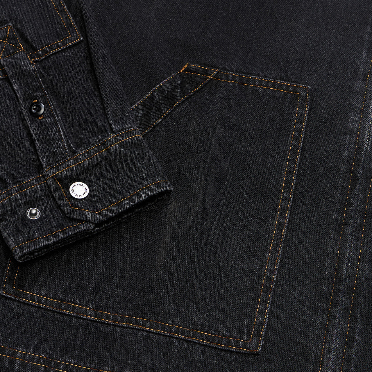 Close-up of a dark washed denim garment showing a large front pocket with contrast tan stitching, a buttoned cuff, and metal snap buttons.