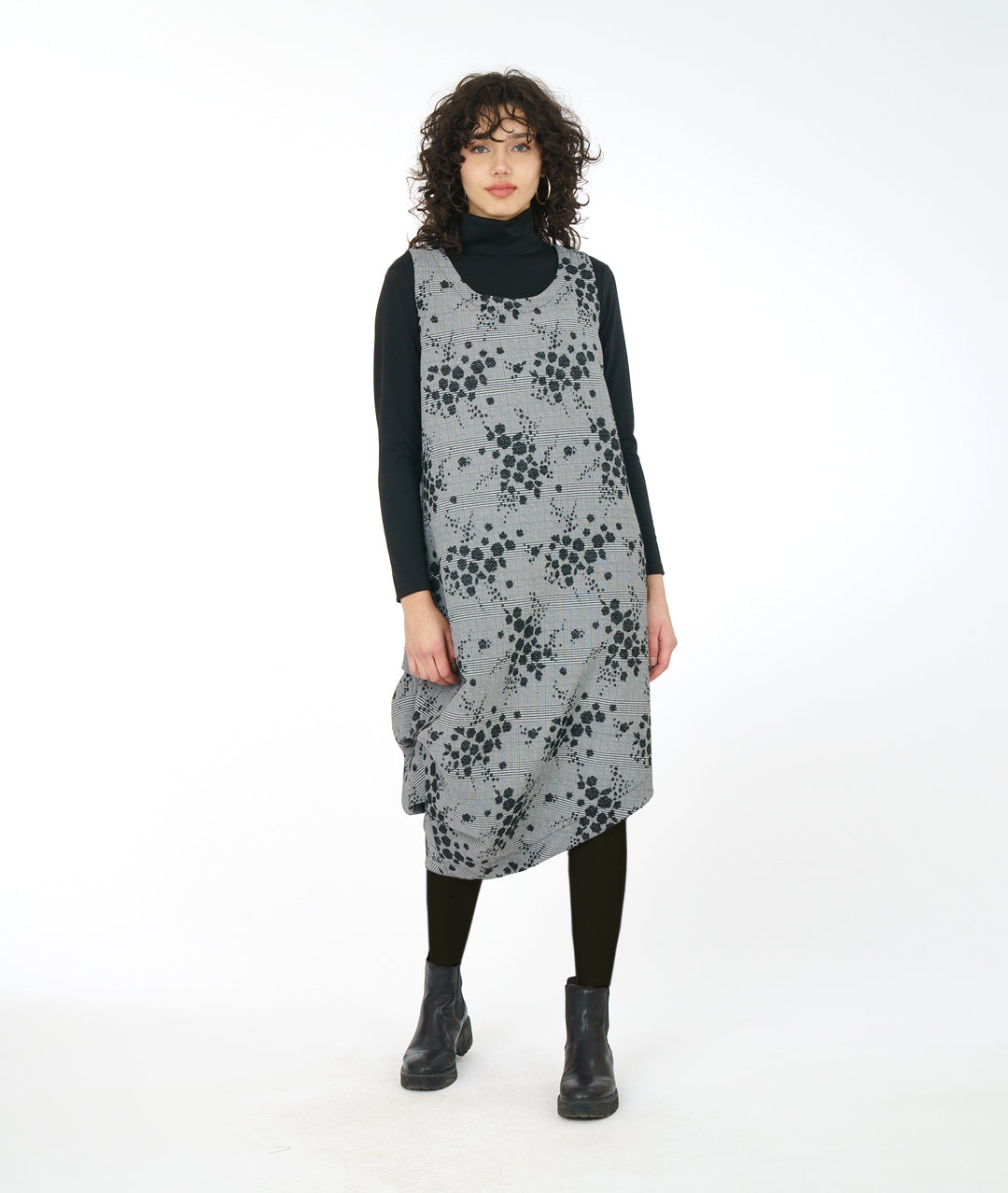 model in a grey plaid asymmetrical jumper dress with a black pollen print, worn over a black turtleneck, leggings and booties