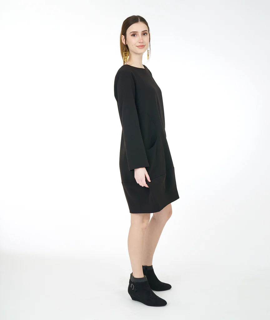 model in a black dress with long sleeves, a wide bottom hem, soft round neckline and curved side seams with a set in pocket on either side, worn with black shoes.