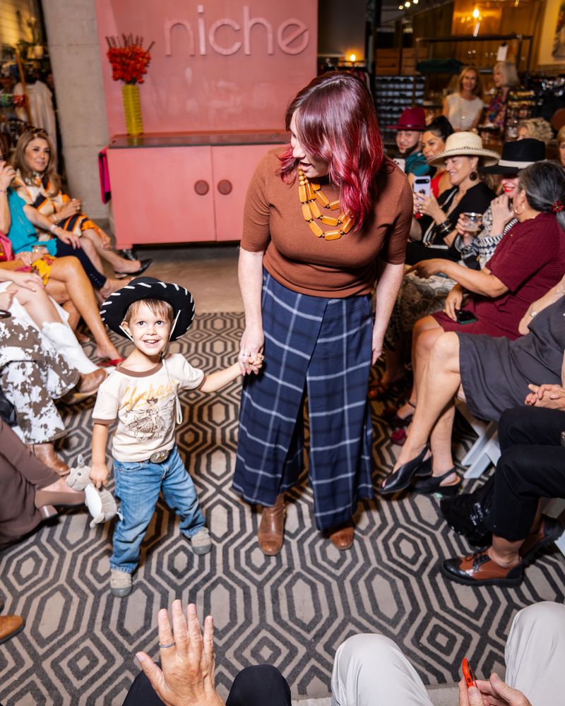 woman wearing brown knit top with layered necklace, navy plaid pants and boots holding hand of little boy wearing black cowboy hat, "yeehaw" shirt and jeans on a runway