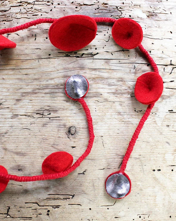 Red felt garland with red yarn strands and silver circular embellishments, laid out on a rustic light wood surface.